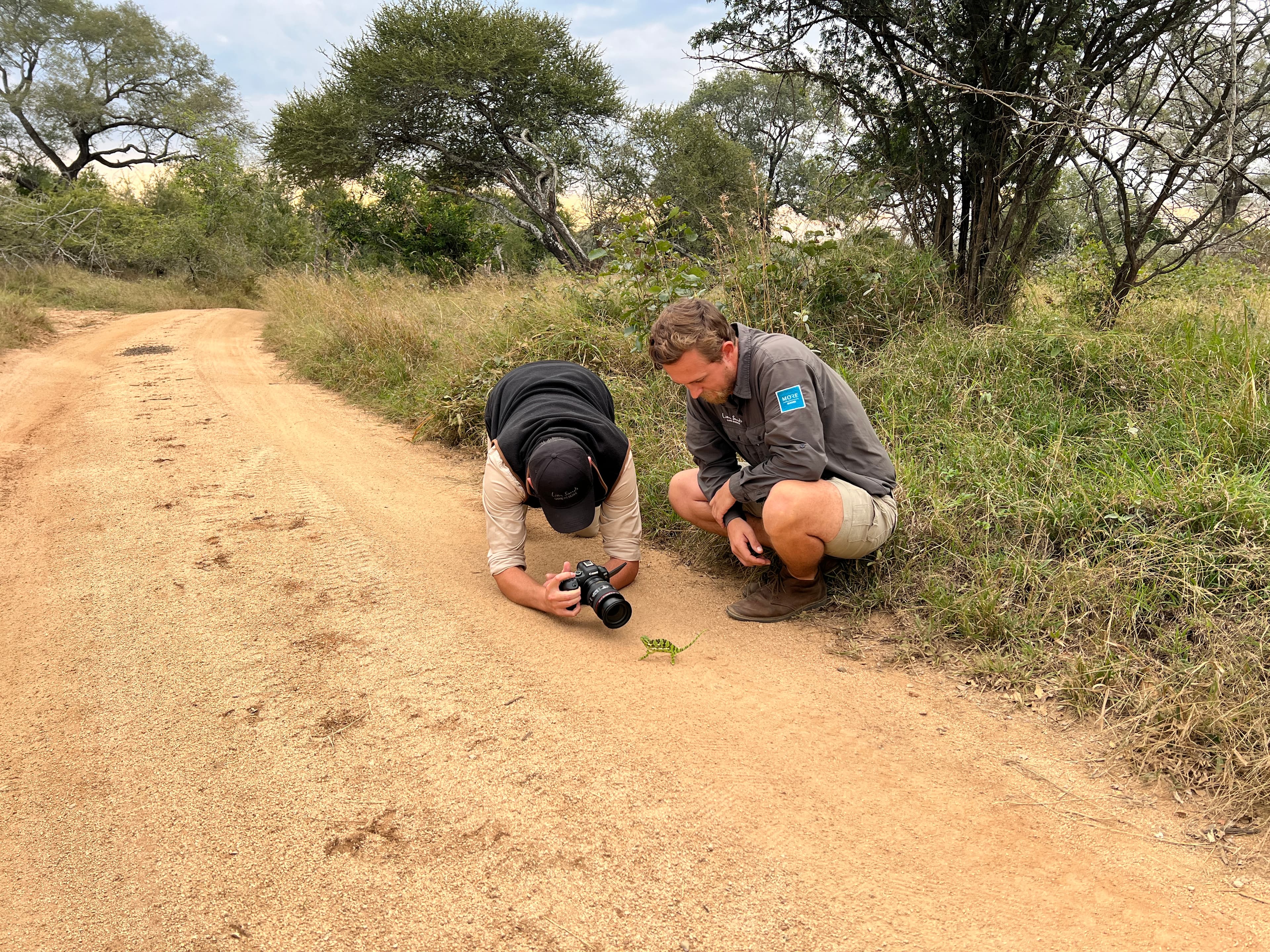 Wildlife photographer crouching on a dirt road with a DSLR, guide alongside